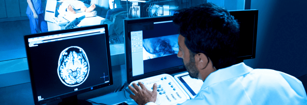 Implementar a Telerradiologia - Médico olhando telas com imagens de exames enquanto pacinete realiza o exame ao fundo